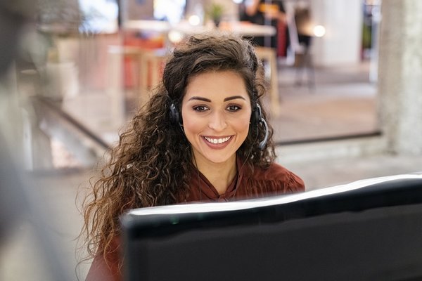 Eine Tourismuskauffrau sitzt mit einem Headset auf dem Kopf an einem Computer und lächelt.